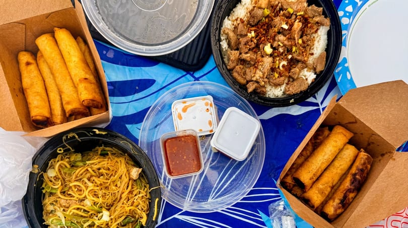 An overhead view of various takeout containers filled with Filipino food, including lumpia, noodles, and rice with meat.