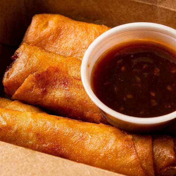 Golden-brown spring rolls and a small cup of dipping sauce in a brown takeout box.